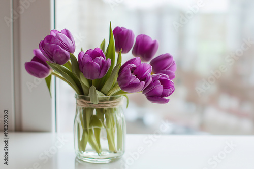 A close-up of a glass vase filled with vibrant purple tulips, placed on a white windowsill with a blurred cityscape in the background.