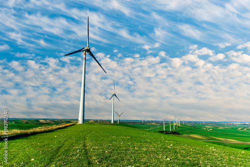 Fotografi wind turbines of wind power plant producing electricity on green sunset hills and cloudy sky on background of landscape