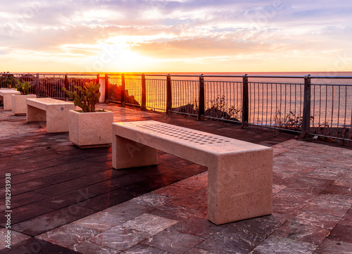 beautiful landscape with white stone bench on seafront embarkment during beautiful sunrise or sunset with sidewalk with pavement, sea surf and nice cloudy sunset sky