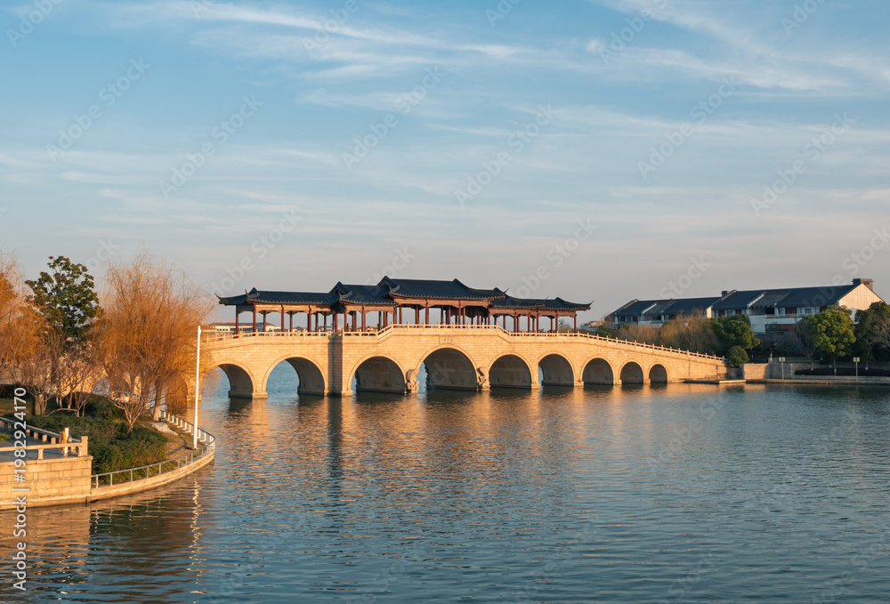Obraz premium Covered stone arch bridge with multiple spans extends across a placid lake during sunset.