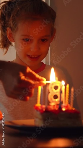 Happy girl sits with cake with burning candles on birthday and laughs. Fathers hands light candles on daughters birthday. Birthday celebration. Vertical shot