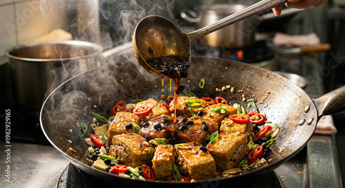 A dynamic action product shot freezing the moment a ladle pours thick, glossy black bean sauce gravy over freshly pan-fried stuffed tofu in a smoking wok. The rectangular tofu pieces, filled with meat