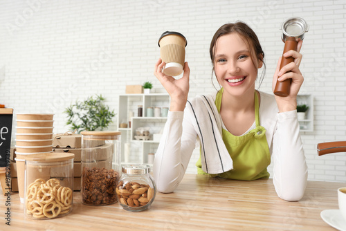 Happy young female barista with paper cup of coffee and portafilter in cafe