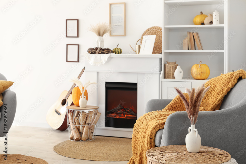 Obraz premium Interior of living room with armchair, coffee table, guitar, autumn home decor on fireplace and pumpkins in shelving unit near white wall