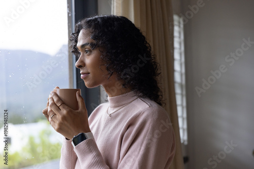 Non-binary adult standing near rain-speckled window holding ceramic mug, wearing pink sweater