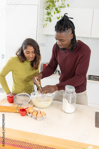 Diverse couple standing in modern home kitchen, whisking batter with metal whisk in glass bowl