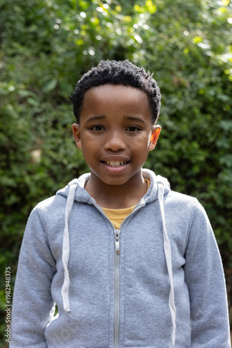 African American child standing by hedge wearing gray hoodie with white drawstrings and yellow tee