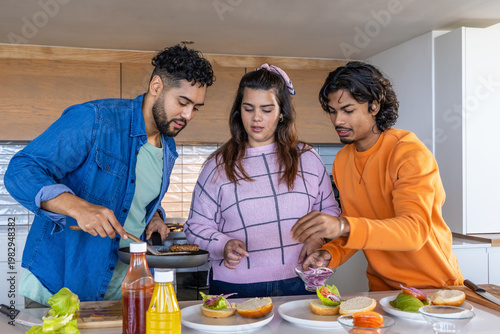 Indian friends cooking burgers at kitchen counter, flipping patties with spatula and topping buns