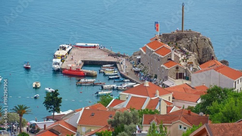 High angle view of Petrovac harbor in Montenegro, featuring traditional orange tiled roofs, a stone pier with boats, and the calm turquoise Adriatic Sea. A perfect summer travel and vacation scene
