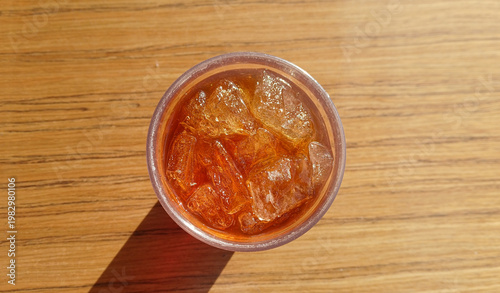 top view of iced tea or black coffee with ice cubes in a glass on a wooden table with sunlight and shadows at a cafe