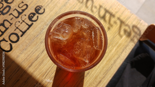 top view of iced tea or black coffee with ice cubes in a glass on a wooden table with sunlight and shadows at a cafe