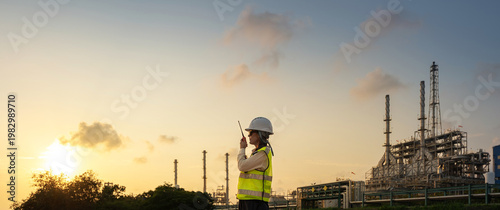 Female petroleum engineer in safety helmet and vest using radio communication for talking project with tablet at oil refinery site during sunset engineering operation and energy industry.