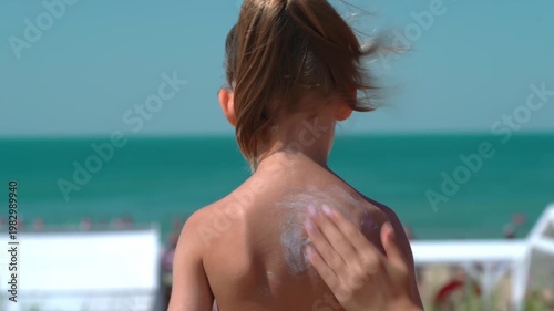 Mothers hands apply protective sunscreen or sunblock on childs skin back on beach with blue sea. Skin care from sun. Summer vacation travel holiday