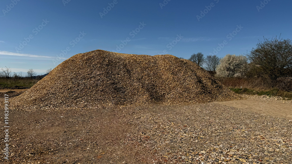 Fototapeta premium Large mound of gravel and stones on a construction site under a clear blue sky with sparse vegetation and trees in the background