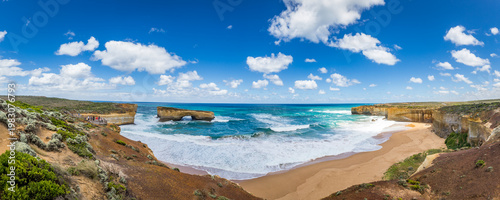 Surreal cliffs and rock formation London Bridge rise from the vibrant ocean, with waves crashing on sandy shores at Great Ocean Road, Victoria, Australia.