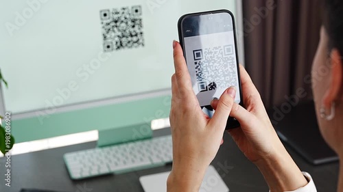 Person scanning QR code from computer screen with smartphone at home workspace. Digital interaction between devices and online service access concept.