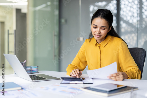 Young professional woman or accountant sitting at an office desk, analyzing financial documents and using a calculator while working with a laptop and other paperwork, managing business finances