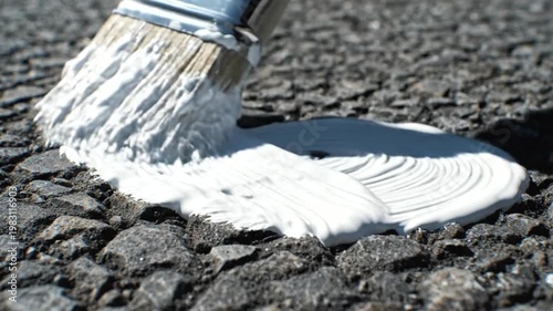 Close up of a paintbrush applying white paint to asphalt surface.