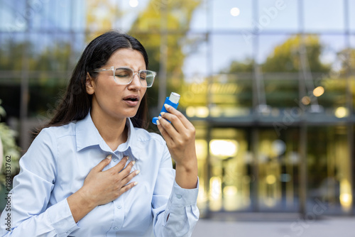 Indian businesswoman clutching an inhaler and pressing her chest during a sudden asthma attack outside a modern glass office building, struggling to breathe and distressed