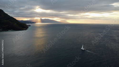 Sailing yacht crossing calm sea at sunset near tropical coastline. Dramatic sun rays break through heavy clouds above distant mountains creating atmospheric golden reflections on open water.