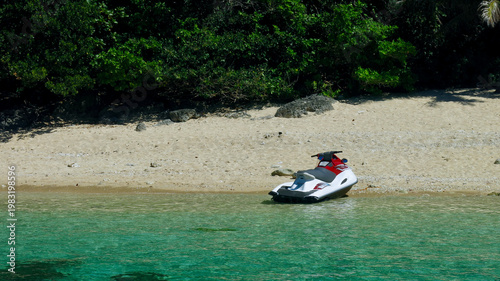 Jet ski parked on tropical sandy beach beside crystal clear water. Personal watercraft rests on secluded shoreline surrounded by turquoise sea and lush coastal jungle.
