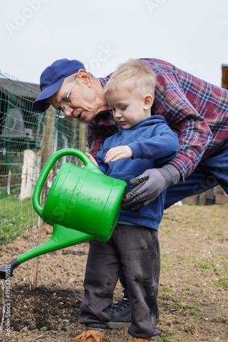 Grandfather helping grandson water plants with watering can in garden, family gardening activity, teaching child to care for nature outdoors