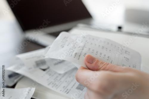 Hand holding receipt with more receipts, notebook and laptop visible in background, personal finance and accounting