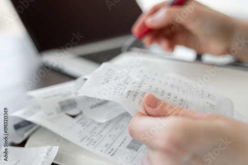 Hand holding receipt with another hand holding pen, more receipts, notebook and laptop visible in background, personal finance and accounting