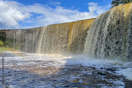 Small but powerful waterfall cascading down on sunny day with light clouds in blue sky, nature and outdoor travel