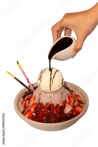 A hand pours dark chocolate syrup over a decadent bowl of chocolate shaved ice topped with ice cream and strawberries.