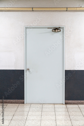 Minimalist light blue industrial door in a hallway with two-tone white and dark grey walls. Features tiled floor, metal doorknob, and overhead door closer in an institutional corridor setting.