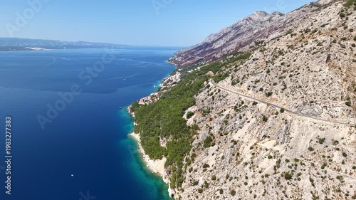 Aerial view of the scenic coastal road winding along the steep rocky mountains of the Dalmatian coast in Croatia, overlooking the turquoise Adriatic Sea on a sunny summer day
