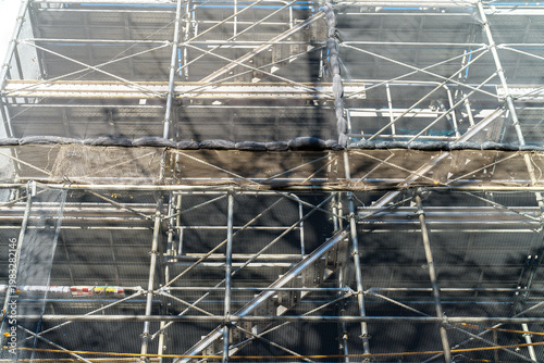 Industrial scaffolding structure with safety netting and metal poles at a construction site. Detailed view of urban building renovation and maintenance equipment in bright daylight.