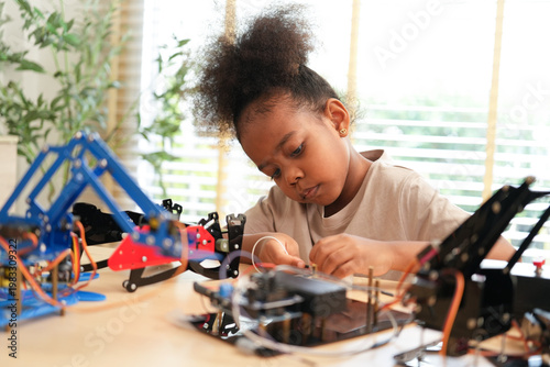 Photos Curious young African American girl concentrates carefully while assembling and wiring a robotic arm kit at the table, exploring STEM learning on her own