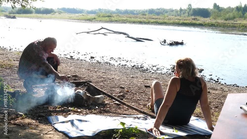 A man and a woman are preparing a barbecue on an open fire near the river.