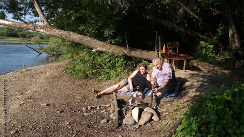 A man and a woman are preparing a barbecue on an open fire near the river.