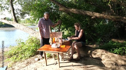 A man and a woman sit on a fallen tree eating and talking during a long outdoor picnic in nature