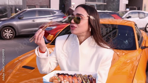Stylish young woman in sunglasses eating sushi with chopsticks while leaning on yellow sports car in city parking lot. Perfect for food, lifestyle, fashion and automotive themed projects
