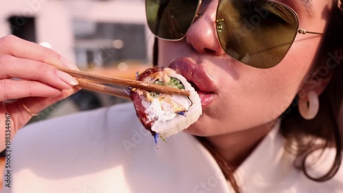 Extreme close-up of young woman in sunglasses eating sushi roll with chopsticks outdoors. Perfect for food, Japanese cuisine, lifestyle, street food and urban themed commercial projects