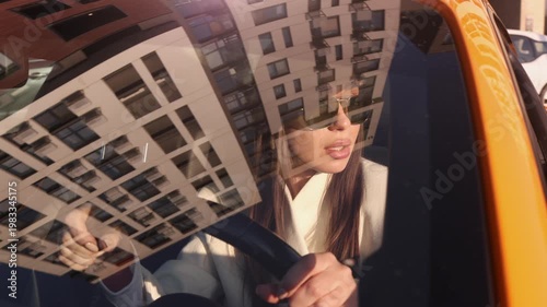 Medium shot of a woman in a white blazer and aviator sunglasses driving an orange car, seen through a window with a strong architectural reflection of a modern apartment building