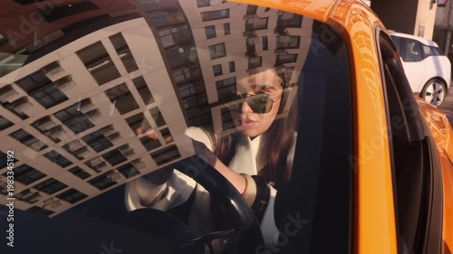 Stylish young woman in sunglasses driving bright yellow car on city street with urban building reflections on windshield. Perfect for automotive, lifestyle and fashion themed projects