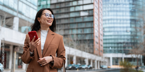 Foto Smiling businesswoman in brown blazer holding cell phone smartphone in modern city street at office buildings, looking away