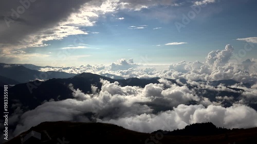 Mountain timelapse above clouds with cabin on alpine ridge at sunrise under dramatic sky. Misty peaks and highland shelter emerge through morning fog beneath glowing dawn horizon.