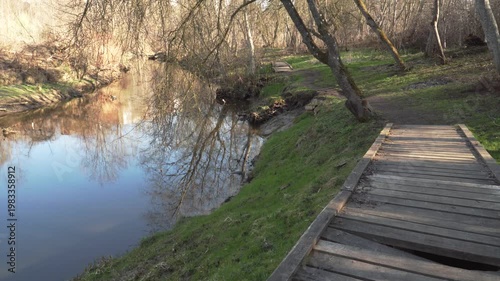 A peaceful river flows next to a weathered wooden boardwalk in the wild. Naked trees and early spring grass line the banks under a bright sun, creating a serene, shady woodland landscape. 