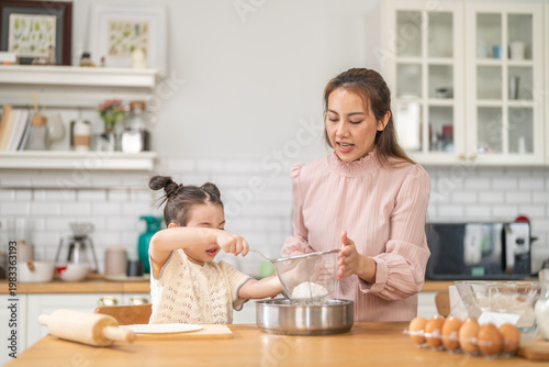 Canvas Print Asian happy mother and little daughter sifting flour with sieve into bowl in kit
