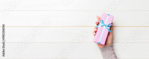 Female's hands holding striped gift box with colored ribbon on white rustic wooden background. Christmas concept or other holiday handmade present box, concept top view with copy space