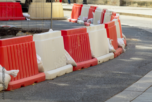 Curved line of red-white barriers with sandbags defines active work zone and directs traffic movement. Scene conveys urban safety measures, controlled access, and temporary infrastructure during stree