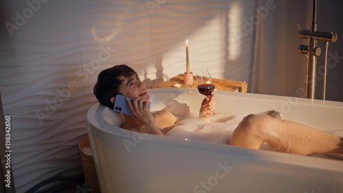 Tableau sur toile Bath girl calling mobile phone lying foam water with red wine glass evening spa