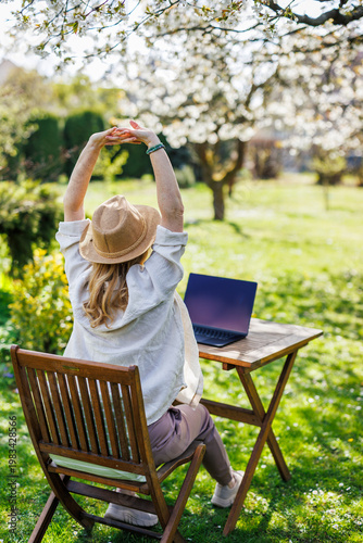 Woman stretching hands while working with laptop outdoors. Stress relief. Exercising body during remote work break in spring garden