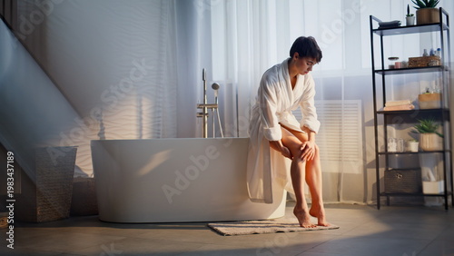 Girl applying legs cream sitting bathtub home. Resting woman rubbing moisturizer